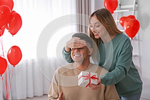 Woman presenting gift to her boyfriend in room decorated with heart shaped balloons. Valentine`s day celebration