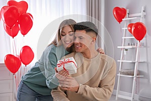 Woman presenting gift to her boyfriend in room decorated with heart shaped balloons. Valentine`s day celebration