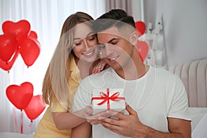 Woman presenting gift to her boyfriend in room decorated with heart shaped balloons. Valentine`s day celebration