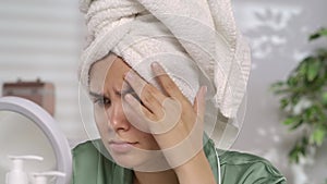A woman with problematic facial skin looks disappointedly in the mirror. A woman after a shower, wearing a white bath