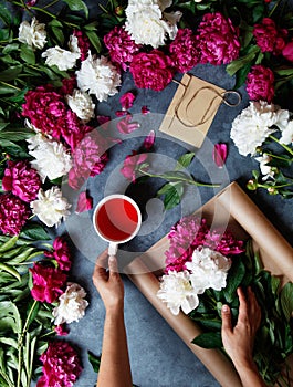 A woman in the process of making a summer bouquet of peonies, holds in her hand a cup of tea karkade. Tools and