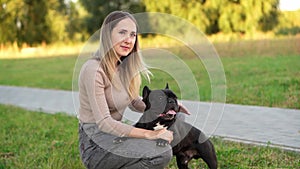 Woman, a professional dog walker, sits down next to a black French bulldog during a walk and pets. Pet care