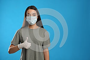 Woman in protective face mask and medical gloves showing thumb up gesture on blue background. Space for text