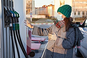 A woman with protective mask chooses a brand of fuel at a gas station for a car. Warm clothes, winter season