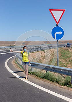 Woman in protective mask hitchhikes on the road on a sunny day.