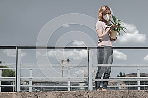 woman in protective mask looking at leaves of potted plant on bridge air