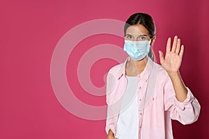 Woman in protective mask showing hello gesture on pink background, space for text. Keeping social distance during coronavirus