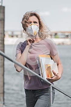 woman in protective mask standing on bridge with books and looking up air