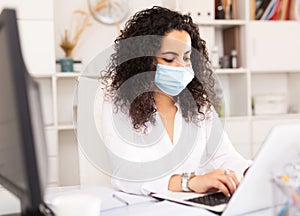 Woman in protective mask worker is working with documents and laptop in office