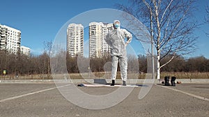 Woman in a protective suit, mask and gloves makes yoga exercises in empty city park.