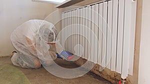 A woman in a protective suit and a respirator sprays a special antifungal spray on the mold-infested wall under the