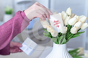 Woman pulling greeting card with Happy mother`s day words from bouquet of white tulips flowers in a white vase on the table.