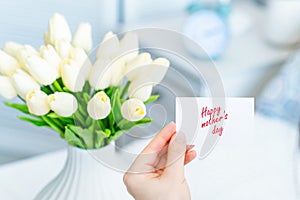 Woman pulling greeting card with Happy mother`s day words from bouquet of white tulips flowers in a white vase on the table.