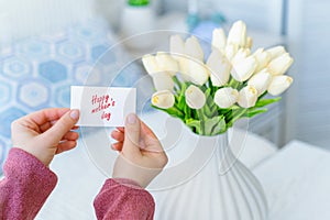 Woman pulling greeting card with Happy mother`s day words from bouquet of white tulips flowers in a white vase on the table.