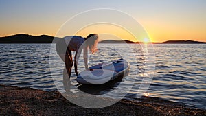 Woman pushes stand up paddle board into water with sun setting in the distance, near Sibenik, on the Adriatic coast of Croatia.