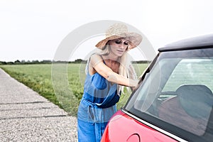 Woman pushing broken down car on country road against clear sky