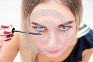 Woman puts black mascara on her eyelashes with makeup, photo of attractive blonde on a white and gray background.