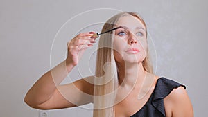 Woman puts black mascara on her eyelashes with makeup, photo of attractive blonde on a white and gray background.