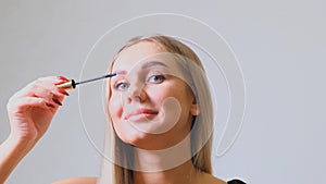 Woman puts black mascara on her eyelashes with makeup, photo of attractive blonde on a white and gray background.