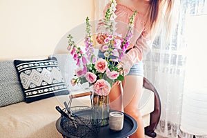 Woman puts bouquet of roses and foxgloves flowers picked in basket in vase at home. Interior and summer decor.