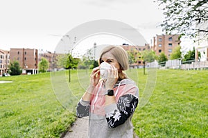 A woman puts on her face mask in a park with buildings in the background