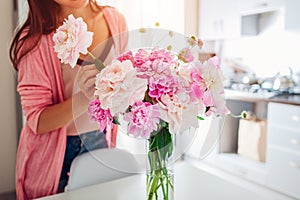 Woman puts peonies flowers in vase. Housewife taking care of coziness and decor on kitchen. Composing bouquet