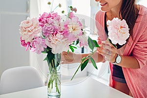 Woman puts peonies flowers in vase. Housewife taking care of coziness and decor on kitchen. Composing bouquet.