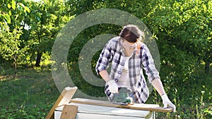 Woman puts on protective glasses and polishes wooden plank with electric sander