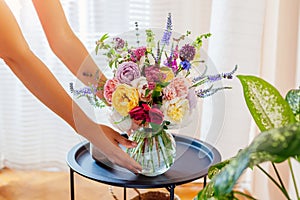 Woman puts transparent vase with bouquet of roses flowers on table. Taking care of interior and summer decor at home
