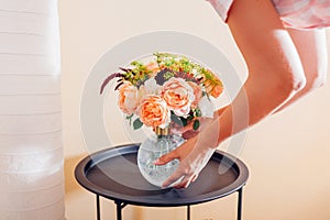 Woman puts vase with bouquet of flowers on table at home. Floral arrangement with orange roses. Interior