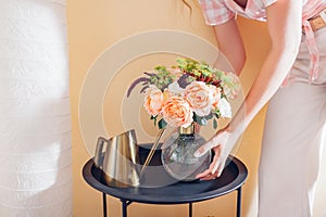 Woman puts vase with bouquet of flowers on table at home. Floral arrangement with orange roses. Interior