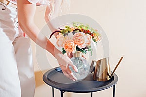 Woman puts vase with bouquet of flowers on table at home. Floral arrangement with orange roses. Interior