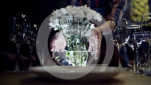 Woman puts vase of flowers on table. Action. Close-up of woman puts vase of white flowers on serving table with