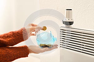 Woman putting coin into piggy bank near thermostat on calorifer. Heating saving concept