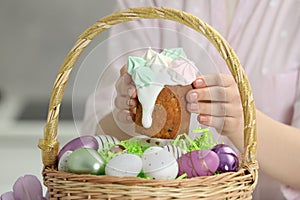 Woman putting delicious Easter cake with meringues into wicker basket with colorful eggs, closeup