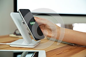 Woman putting mobile phone onto wireless charger at table, closeup. Modern workplace accessory