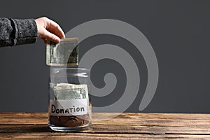 Woman putting money into donation jar on wooden table against grey background. Space for text