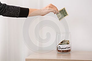 Woman putting money into jar with label DONATION on table against light background