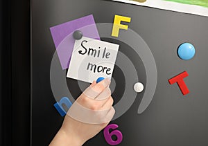 Woman putting note with phrase SMILE MORE on refrigerator door