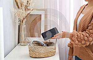 Woman putting smartphone into wicker basket with gadgets at home. Digital detox concept