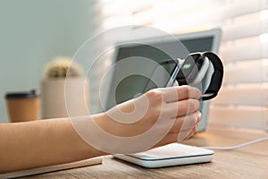 Woman putting smartwatch onto wireless charger at table, closeup. Modern workplace accessory