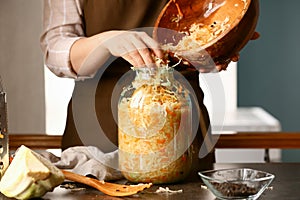 Woman putting tasty sauerkraut into glass jar on table in kitchen, closeup