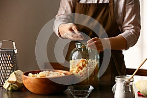 Woman putting tasty sauerkraut into glass jar on table in kitchen, closeup