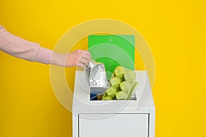 Woman putting used foil container into trash bin on color background, closeup