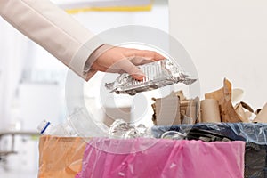 Woman putting used foil container into trash bin in office. Waste recycling