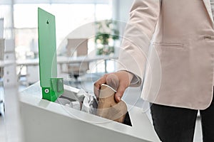 Woman putting used paper cup into trash bin in modern office. Waste recycling