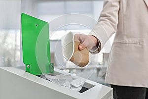 Woman putting used paper cup into trash bin in modern office. Waste recycling