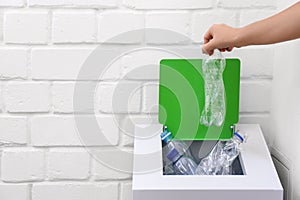 Woman putting used plastic bottle into trash bin near white brick wall, closeup with space for text. Recycling problem