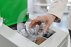 Woman putting used plastic bottle into trash bin in office, closeup. Waste recycling