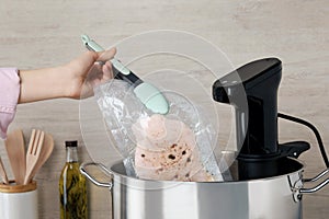 Woman putting vacuum packed meat into pot with sous vide cooker, closeup. Thermal immersion circulator